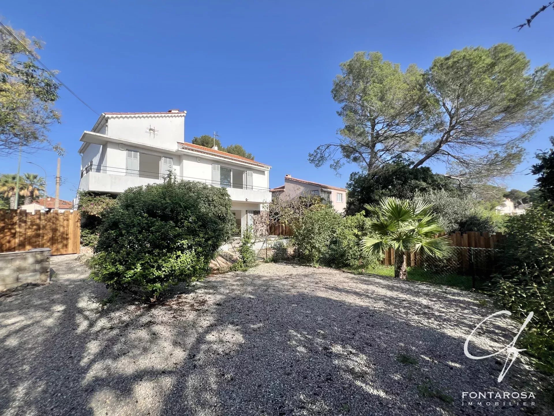 Une maison blanche avec un toit en tuiles se dresse au milieu de la verdure, sous un ciel bleu dégagé.