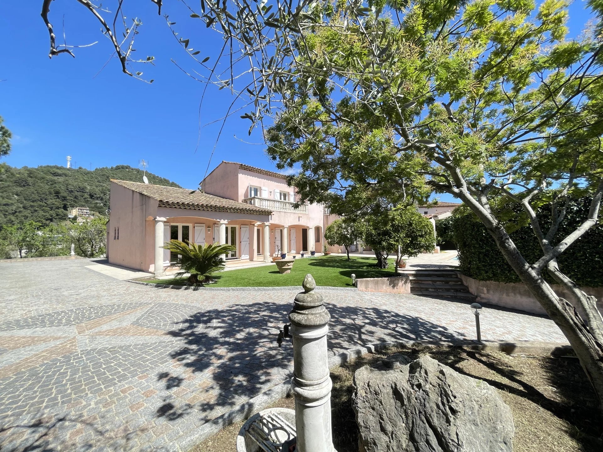 A charming house with a tiled roof is surrounded by trees and a cobblestone driveway under a clear blue sky.