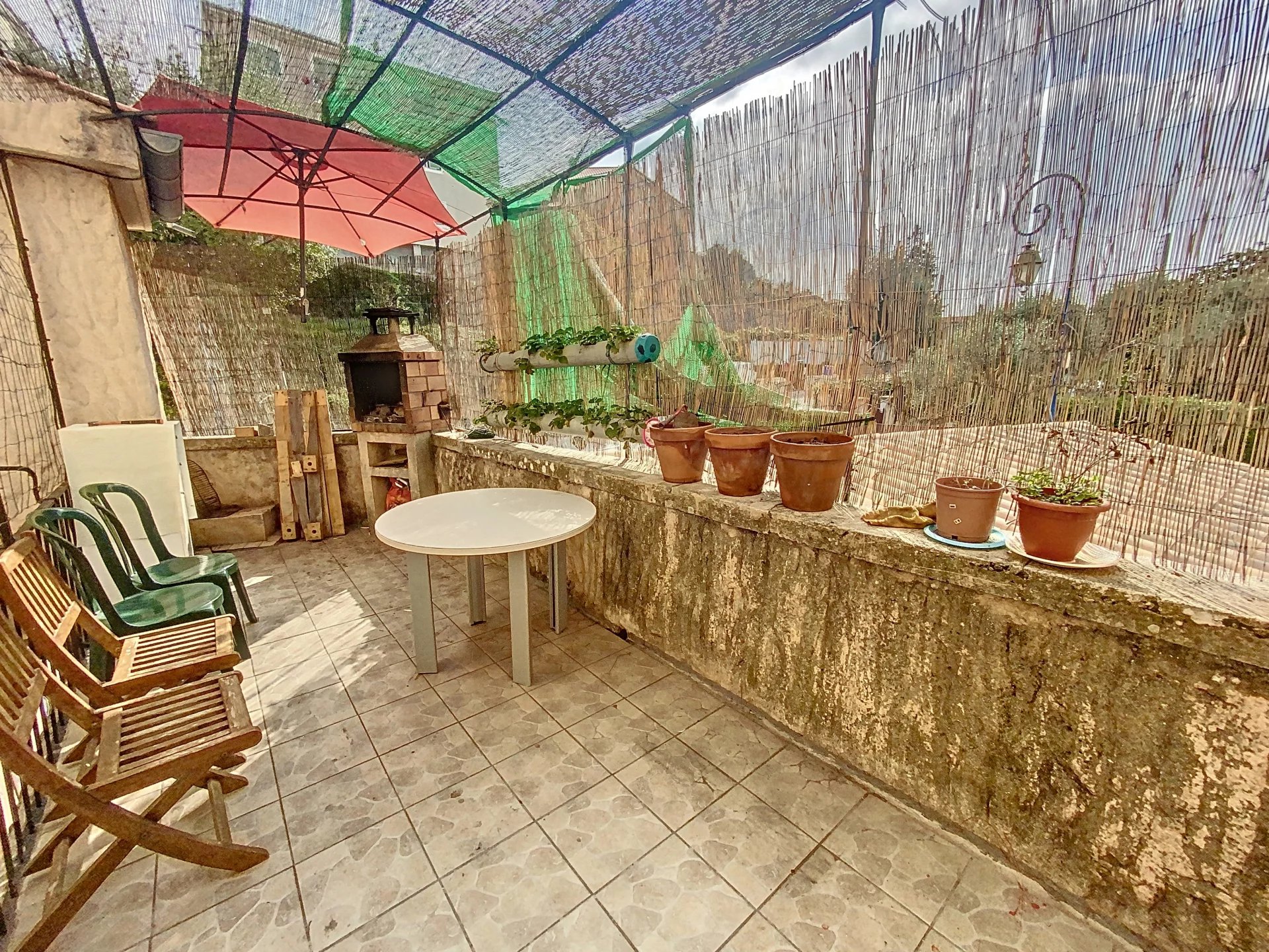 A cozy outdoor patio with a mix of wooden and plastic chairs, a round table, an umbrella, potted plants, and a grill, all under a shaded canopy.