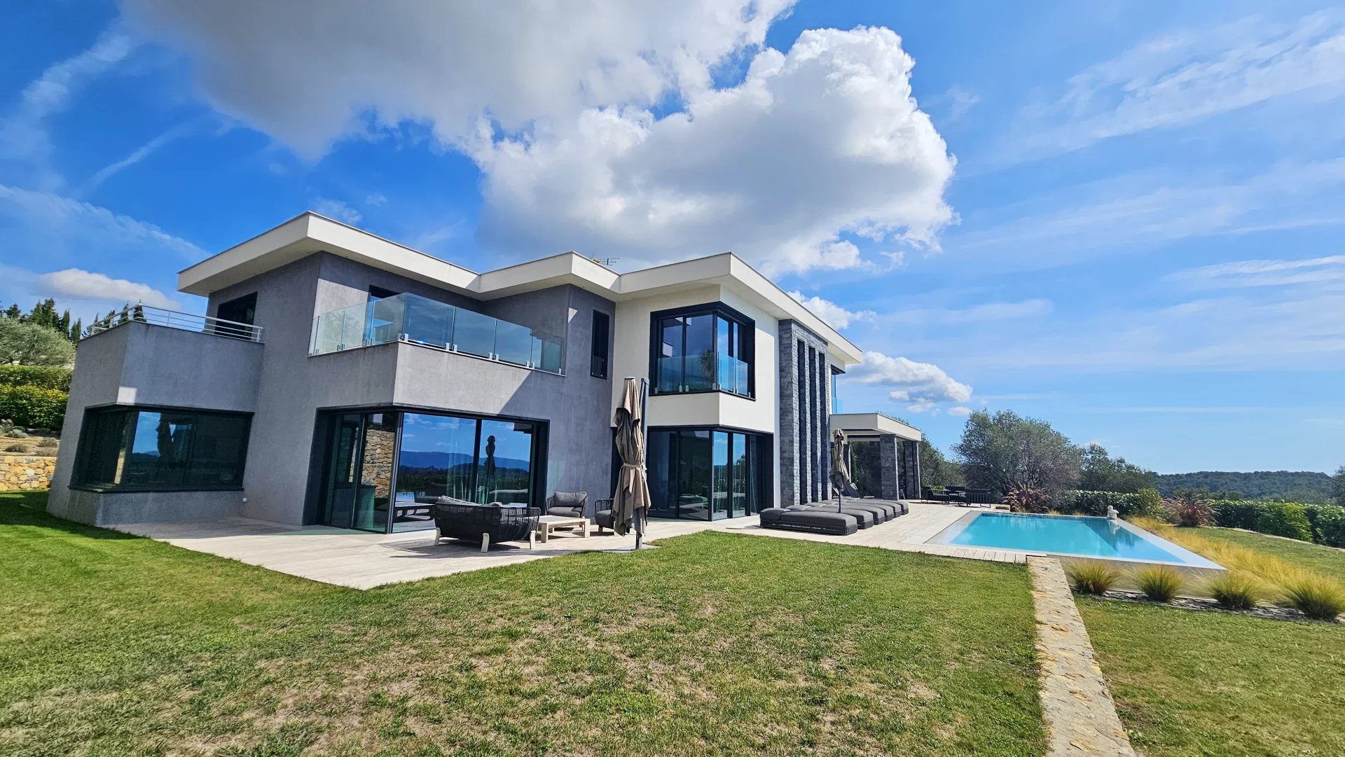 Belle maison moderne avec piscine, entourée de verdure, sous un ciel ensoleillé.