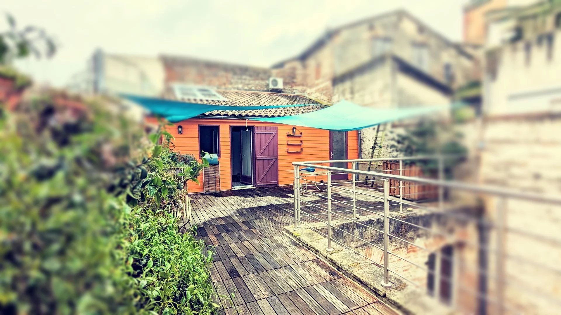 Une terrasse en bois avec un bâtiment en bois orange et une toile d'ombrage bleue, entourée de végétation.