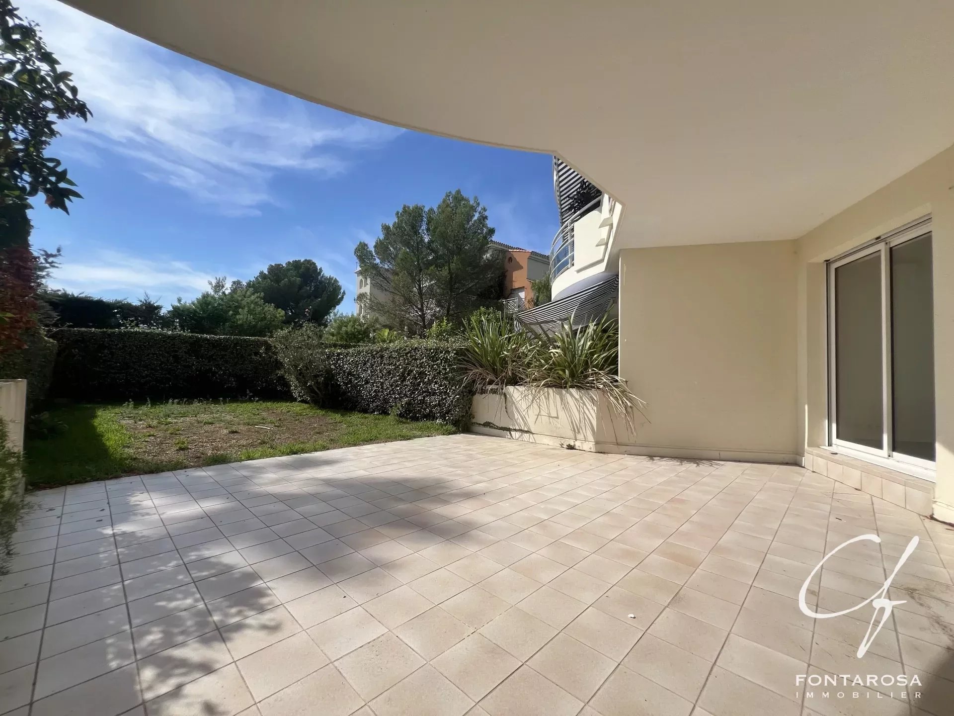 Spacious tiled patio with a small garden area, bordered by hedges and overlooked by residential buildings.