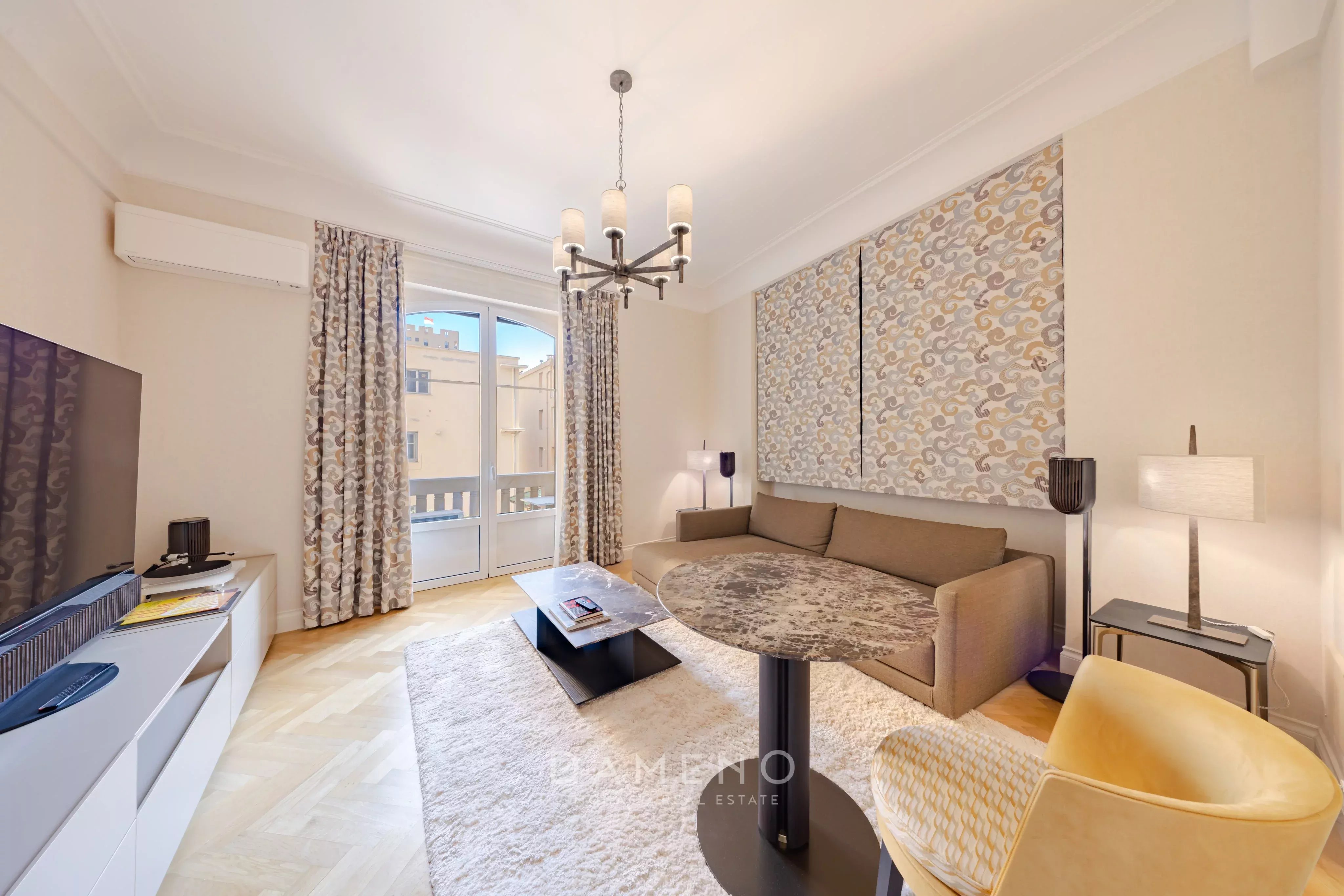 Un salon moderne et lumineux avec un canapé, une télévision, une table basse, et un grand tapis, avec une baie vitrée donnant sur l'extérieur.