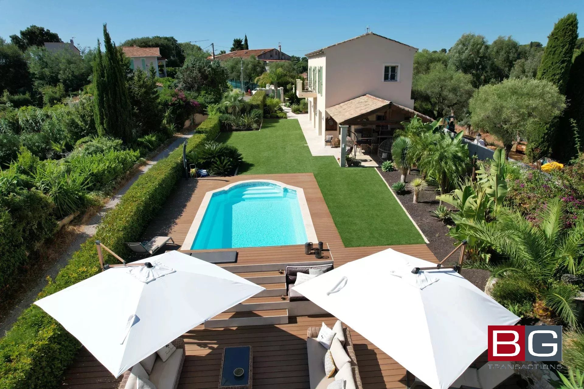 Aerial view of a house with a large garden and a swimming pool. The poolside area features lounge chairs and umbrellas, surrounded by lush greenery.