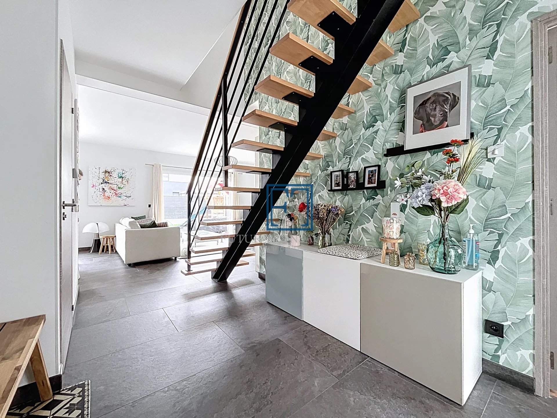 L'image montre un intérieur moderne avec un escalier ouvert en bois et métal, un mur décoré de papier peint à motifs, et un meuble avec des fleurs et des cadres.