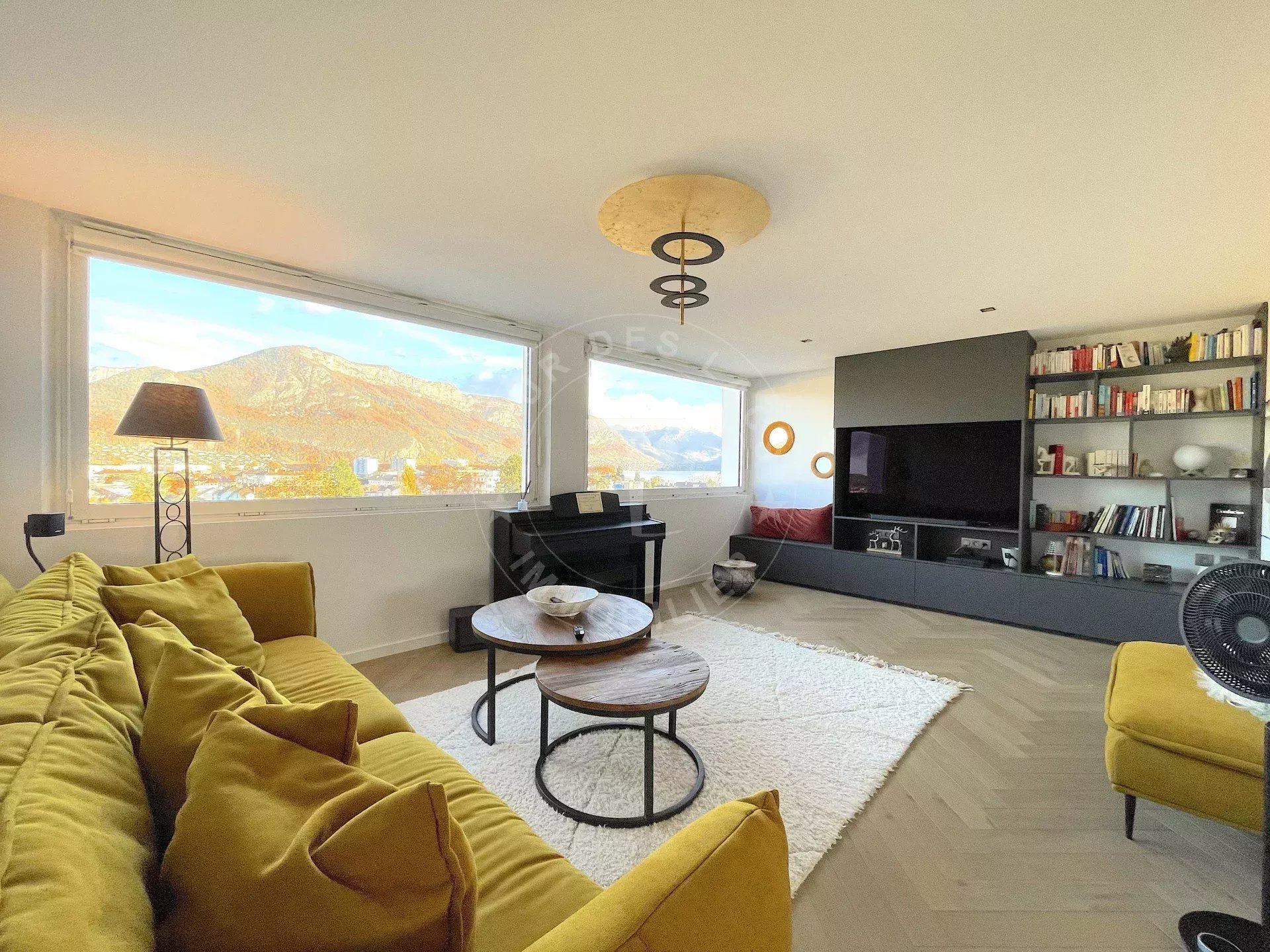 Un salon moderne et lumineux, avec de grandes fenêtres offrant une vue sur les montagnes, un canapé jaune, une table basse, un piano et des étagères avec des livres.