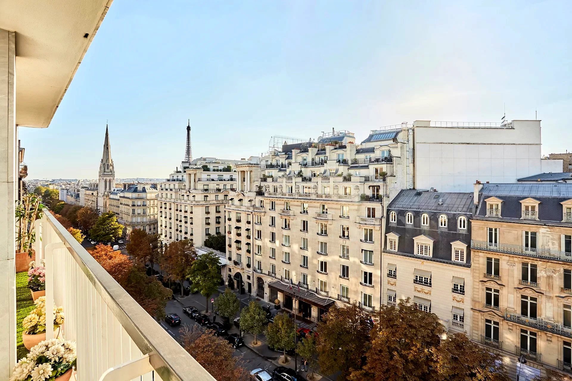 A scenic cityscape view from a balcony, showcasing elegant Parisian buildings, a tree-lined street, and distant landmarks under a clear blue sky.