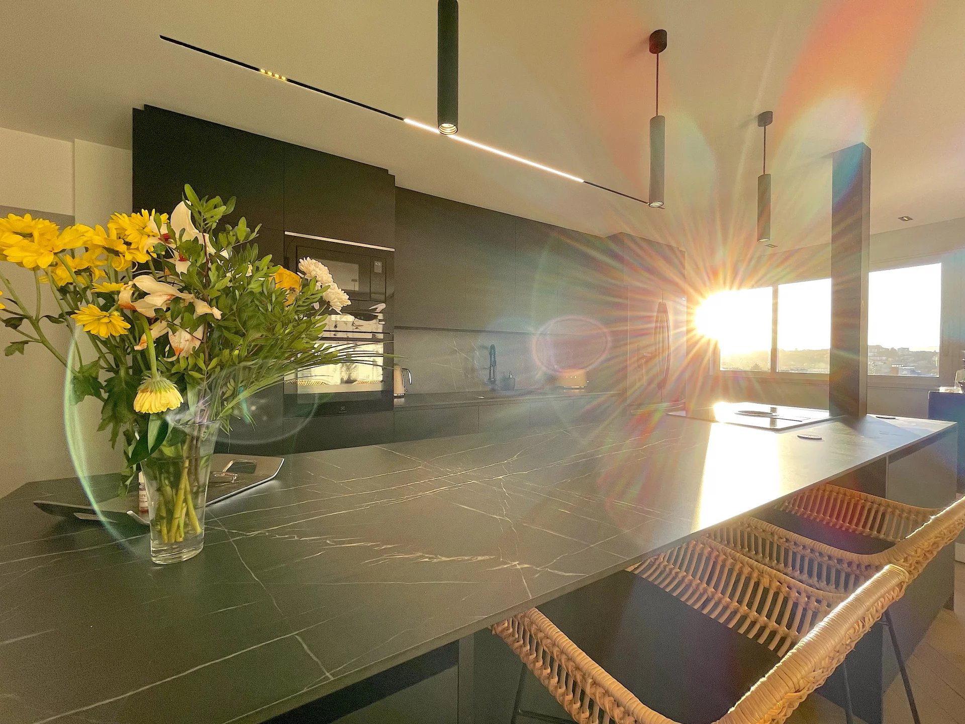 Modern kitchen with a black marble island, wicker chairs, and a vase of yellow flowers. Sunlight streams in through large windows, creating a warm, inviting atmosphere.