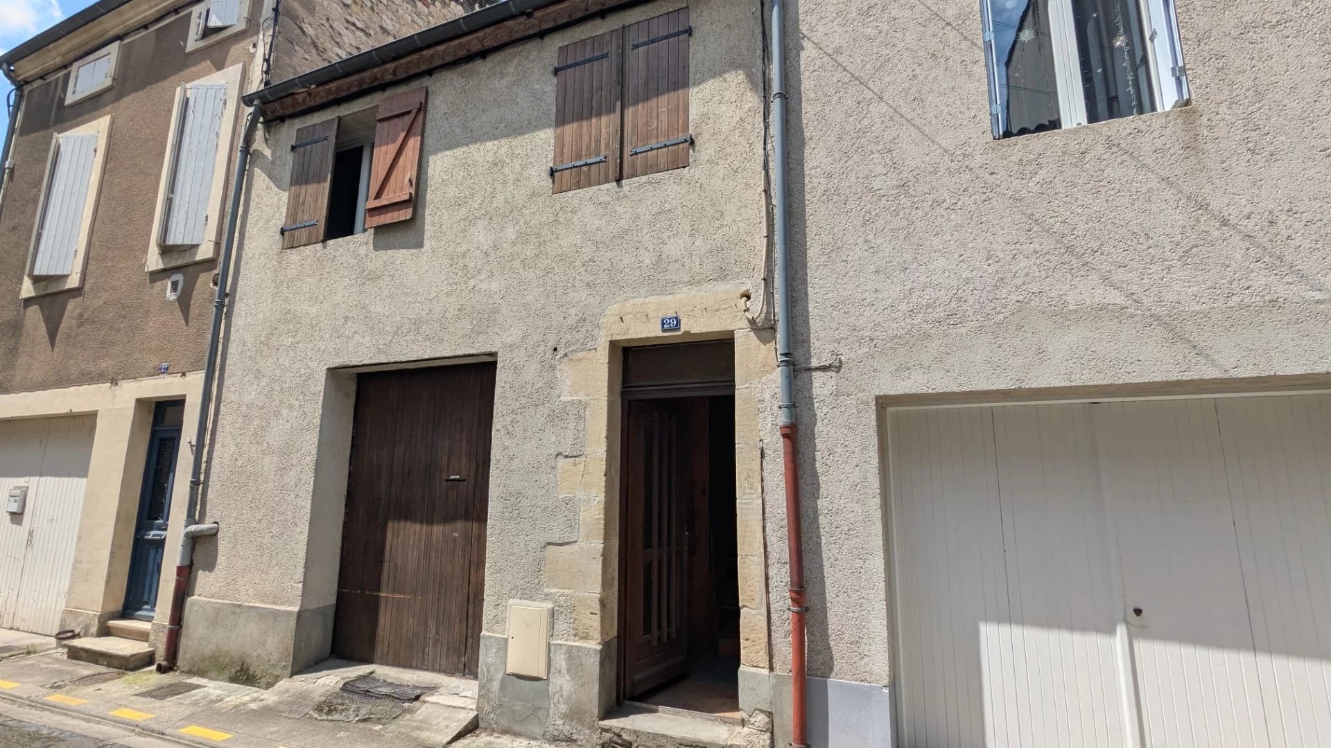 Il s'agit d'une façade de maison en pierre avec des volets en bois et un garage marron, située dans une rue résidentielle.