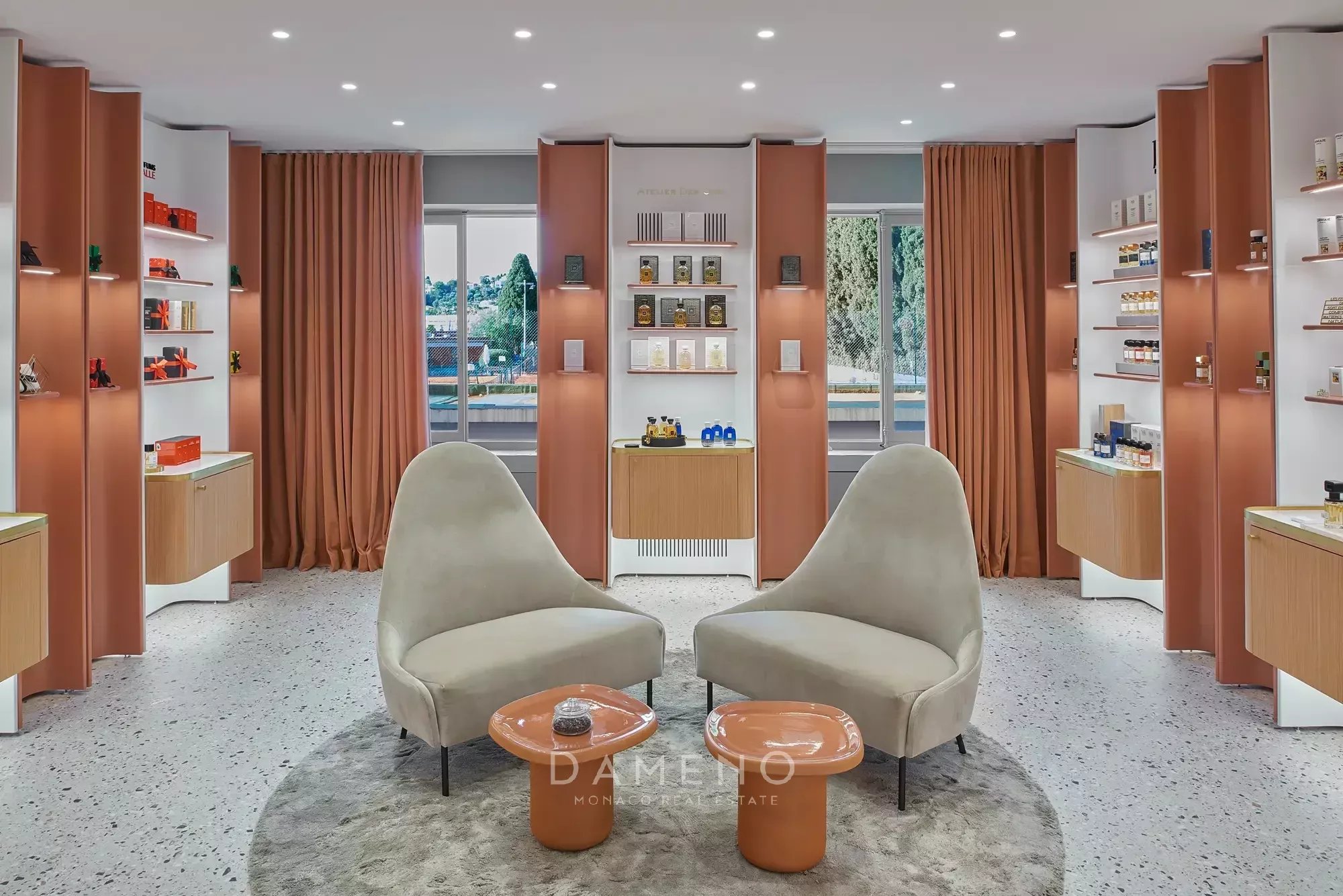 غرفة أنيقة تحتوي على كرسيين مريحين وطاولتين صغيرتين، محاطة بأرفف تعرض منتجات مختلفة، بإضاءة جيدة وستائر برتقالية.