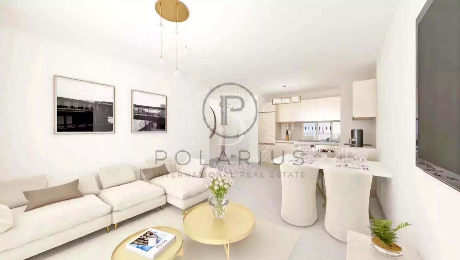 Un salon moderne et lumineux avec canapés beiges, table basse dorée, et une cuisine ouverte en arrière-plan.