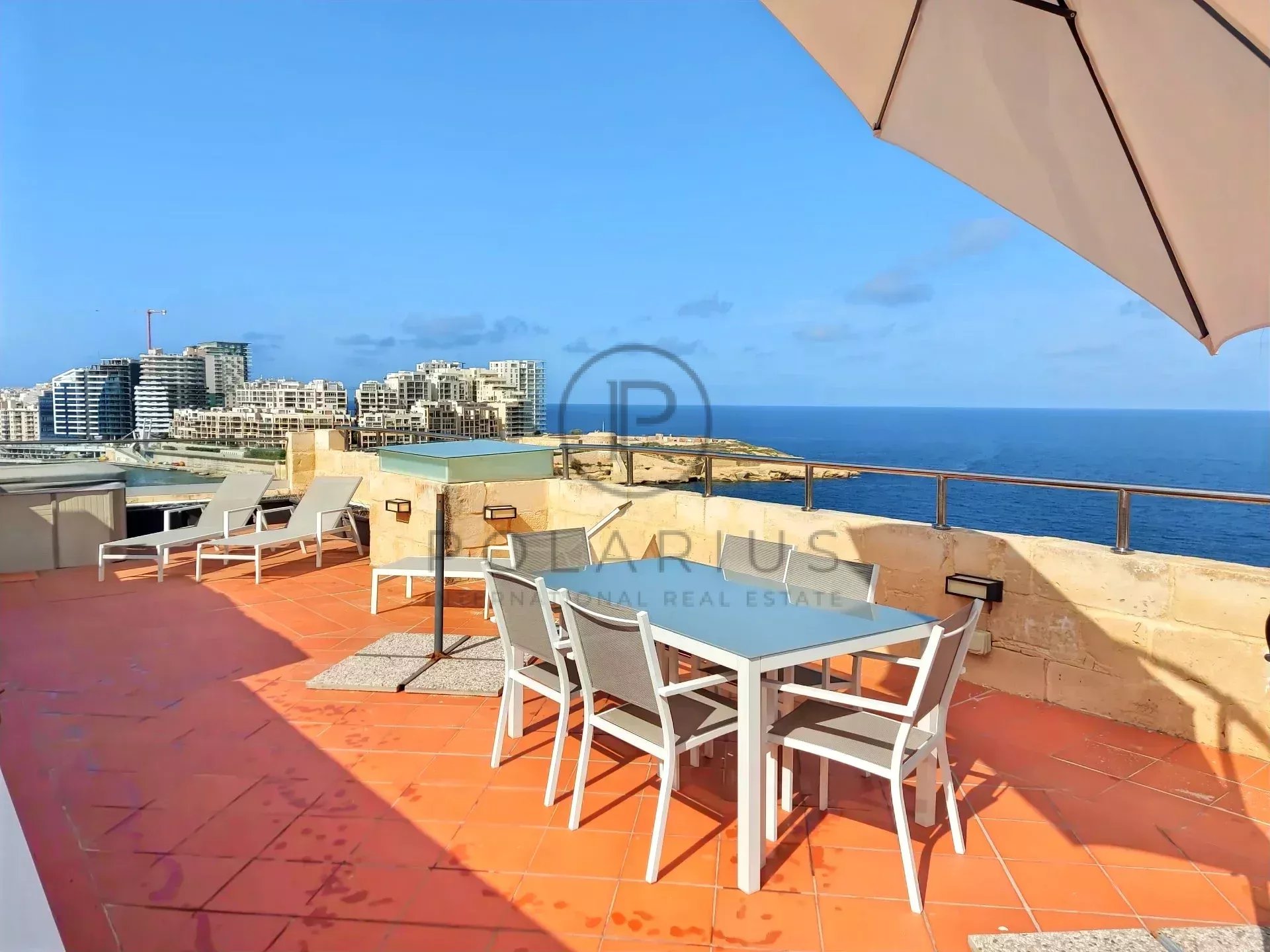 Eine Terrasse mit einem Tisch, Stühlen und Liegen bietet einen Blick auf das Meer und Gebäude in der Ferne, bei klarem Himmel und Sonnenschein.