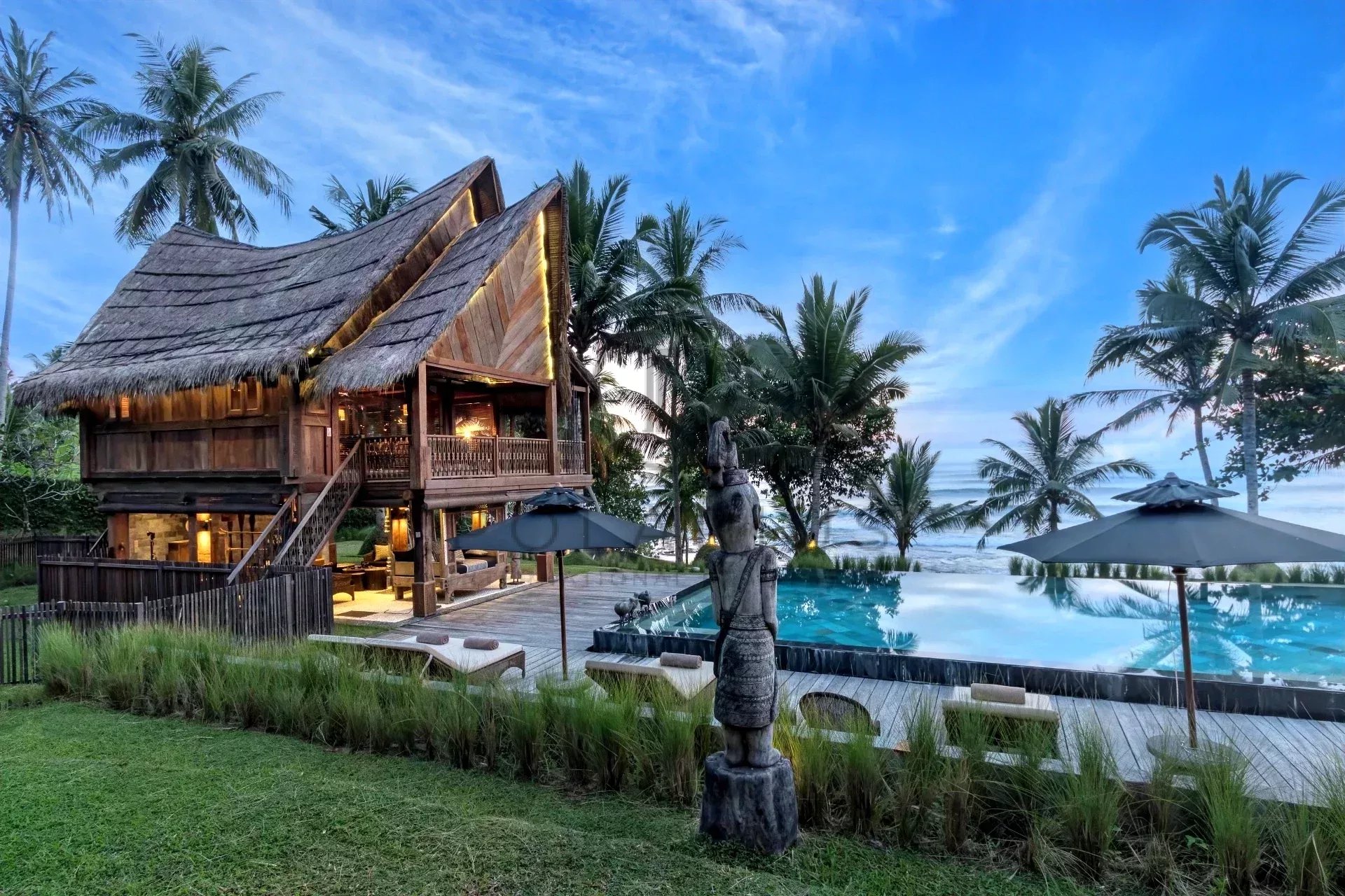 Belle maison en bois avec toit de chaume, située près d'une piscine et entourée de palmiers sous un ciel bleu.
