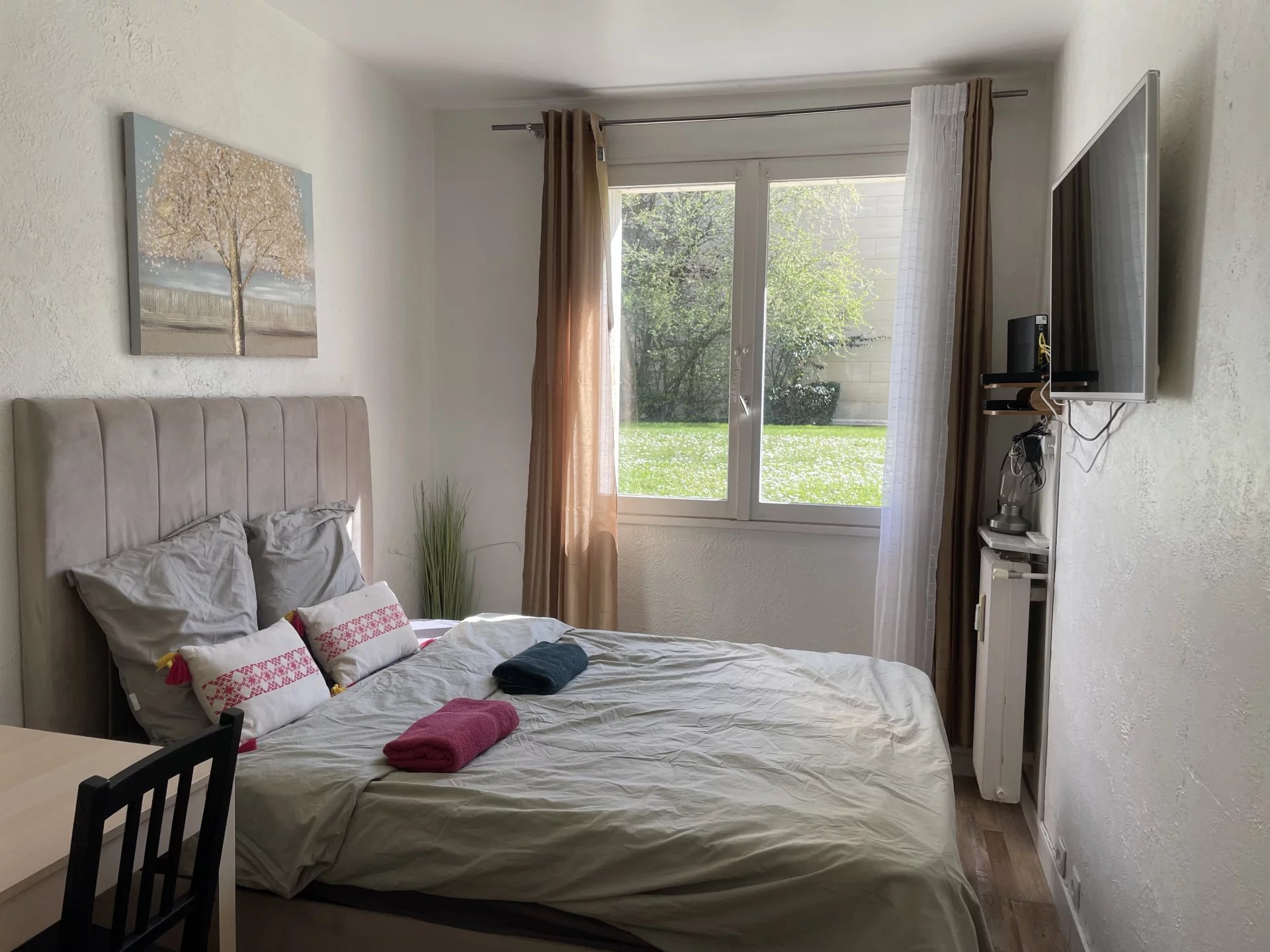 Une chambre moderne avec un lit double, une grande fenêtre, des rideaux beiges, et une télévision murale.