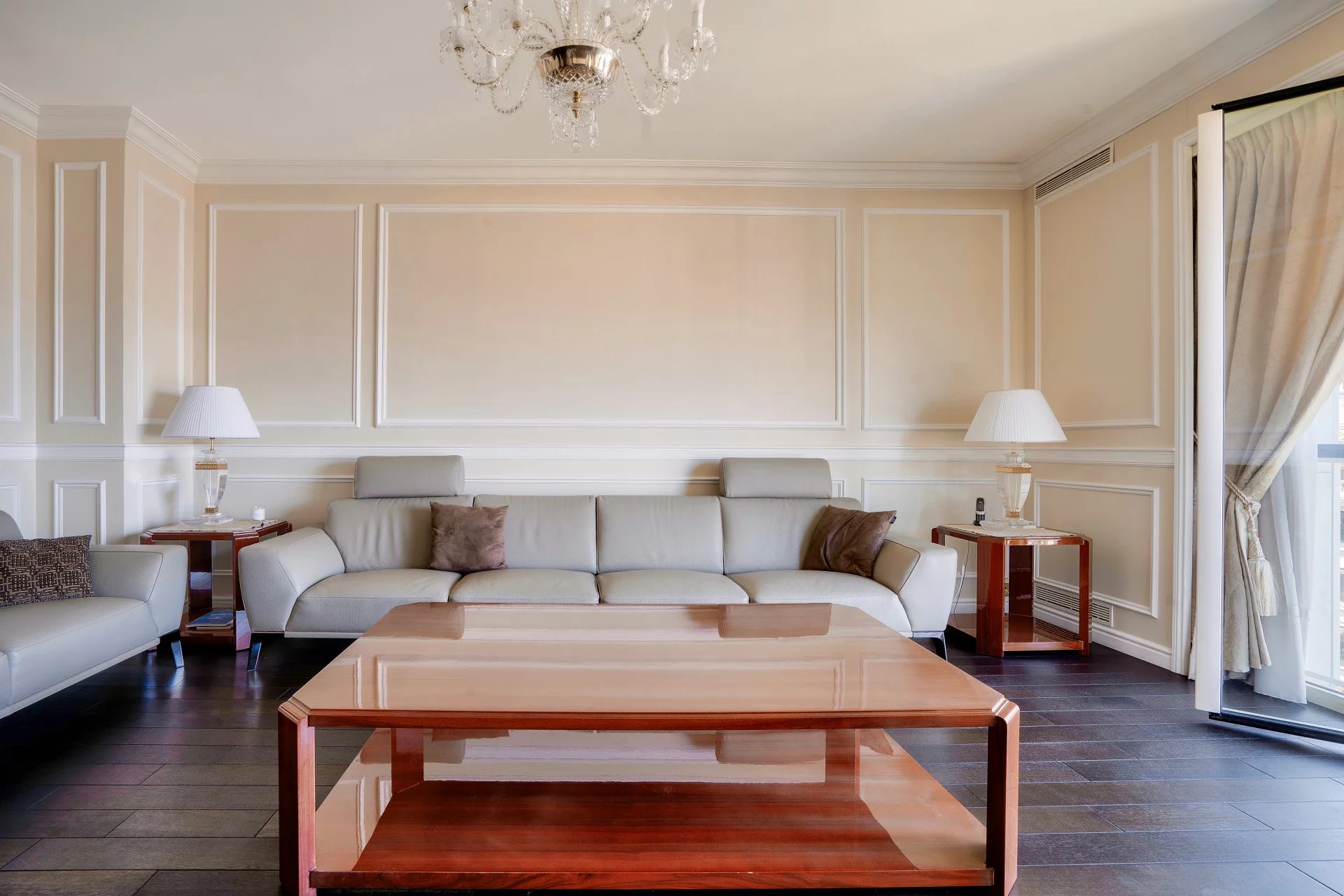 A modern living room with a large beige sofa, wooden coffee table, elegant chandelier, and floor-to-ceiling curtains.