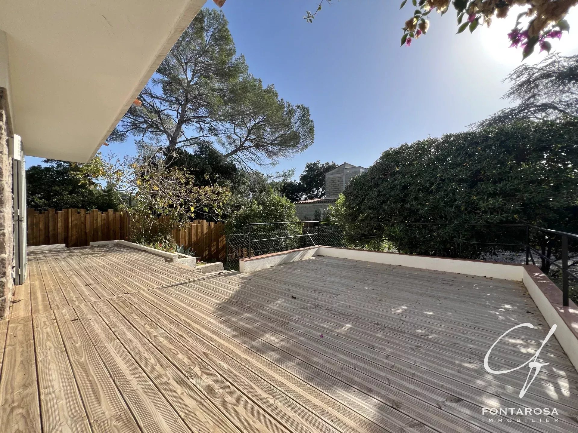 Une terrasse en bois avec vue sur un jardin verdoyant et des arbres sous un ciel bleu.